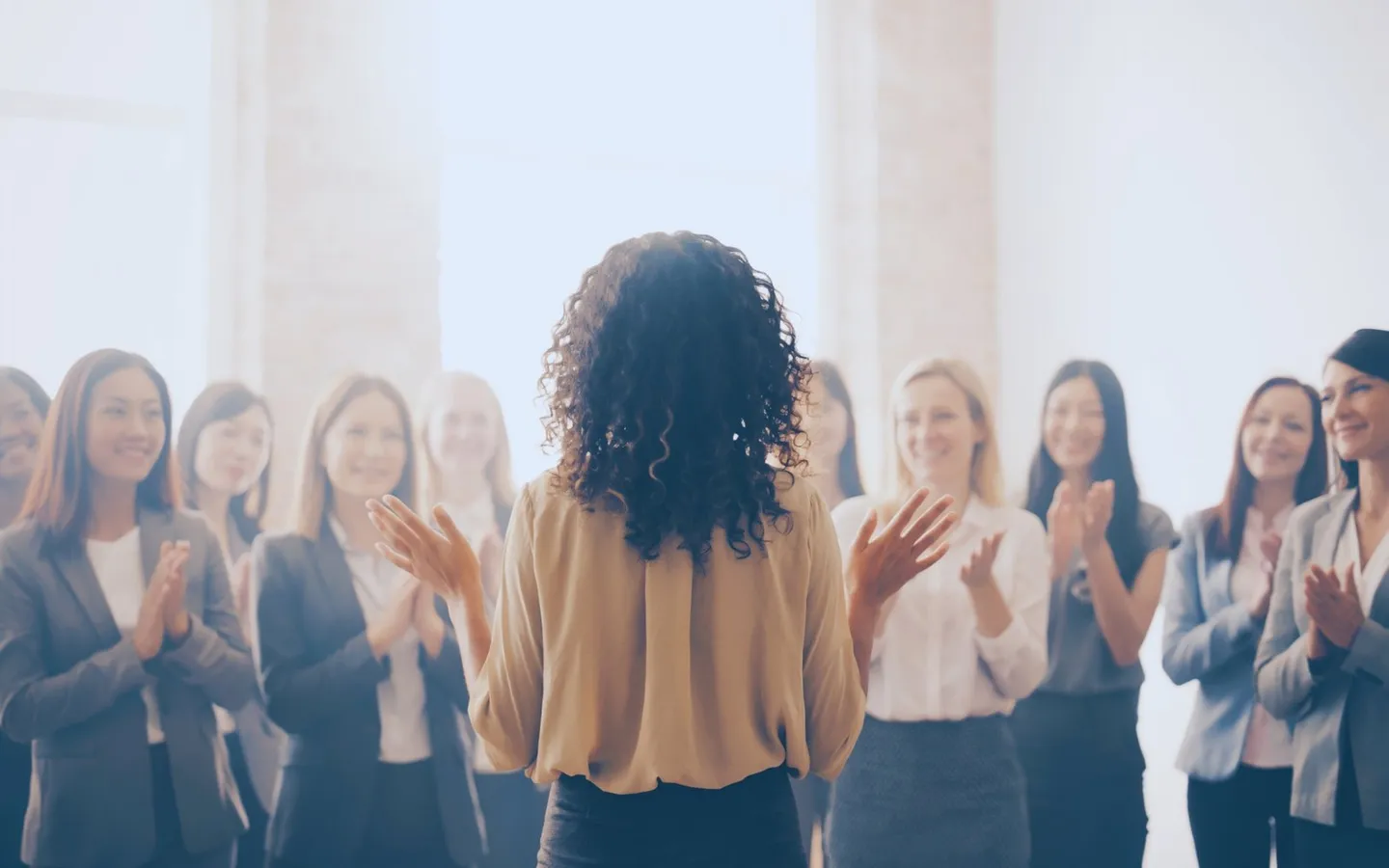 Une femme de dos, debout face à un groupe de collègues qui l'applaudissent dans une salle lumineuse.