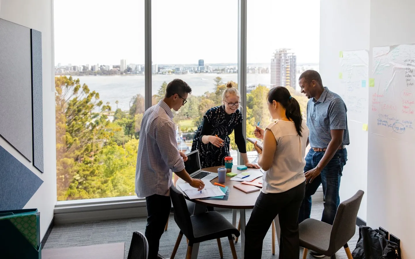 Quatre collègues debout autour d'une table de réunion, devant une grande baie vitrée, en train de travailler ensemble sur des documents.