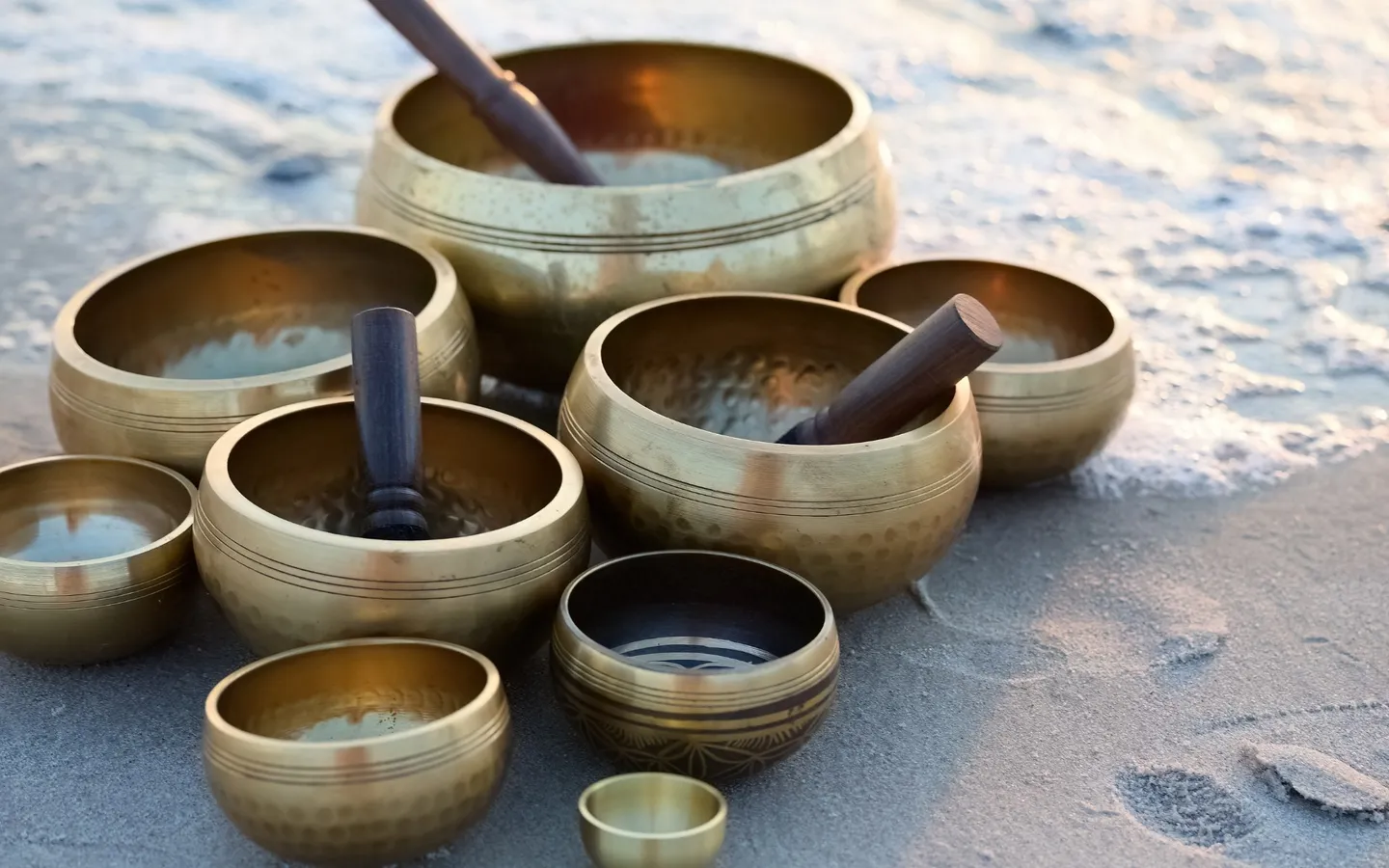 Un ensemble de bols chantants en métal, posés sur le sable au bord de l'eau, avec leurs maillets de bois.