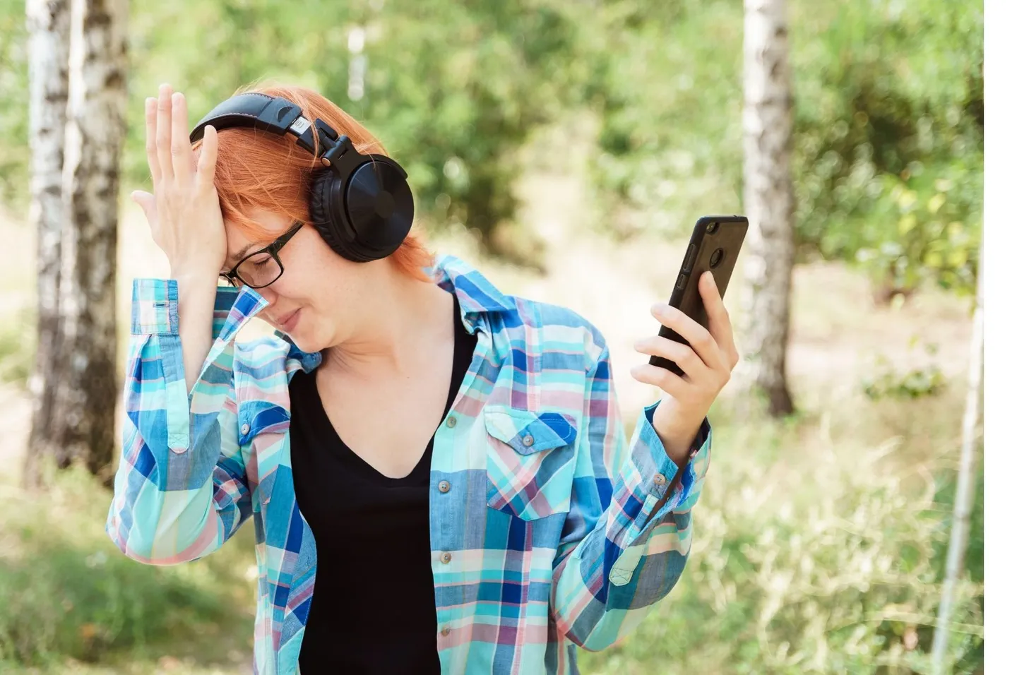 Une femme coiffée d'un casque audio, en extérieur, la main sur le front et les yeux baissés vers son téléphone.