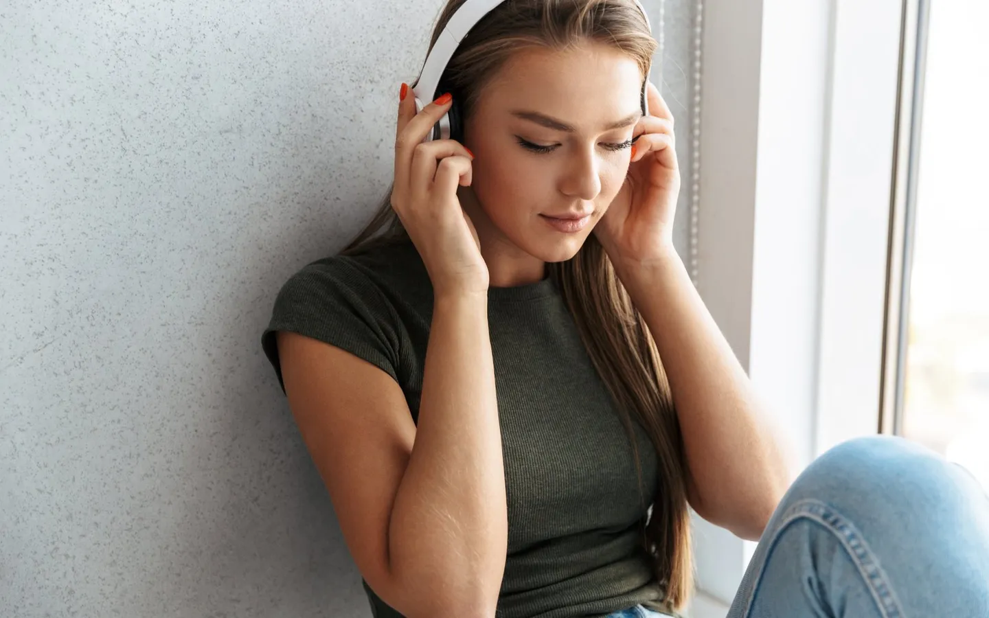 Une jeune femme assise contre un mur, un casque audio sur les oreilles, les yeux baissés.