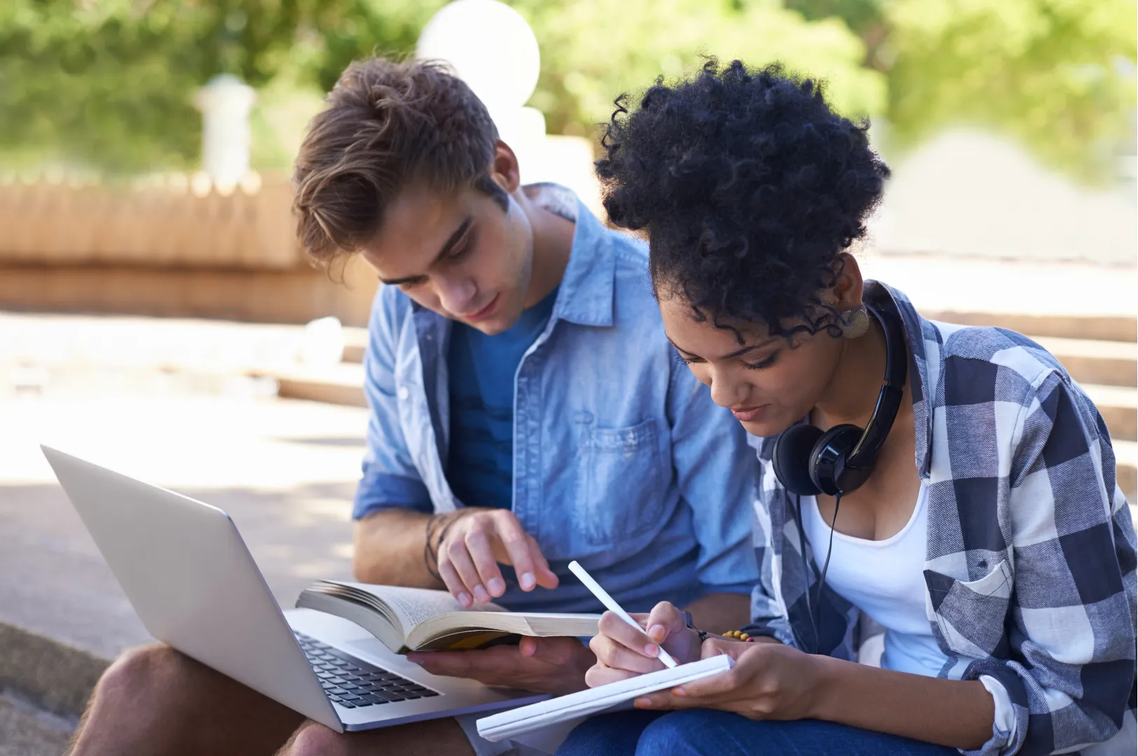 Deux étudiants révisent ensemble à l'extérieur, un ordinateur et un cahier sur les genoux.