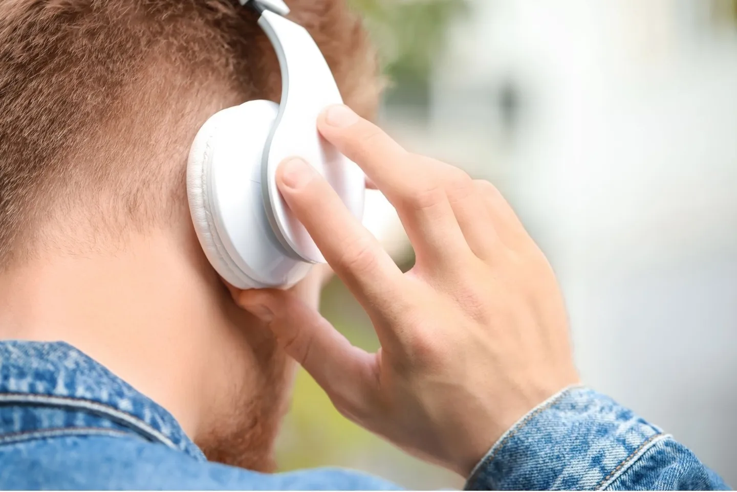 Vu de dos, une main ajuste un casque audio blanc sans fil sur l'oreille, en extérieur.