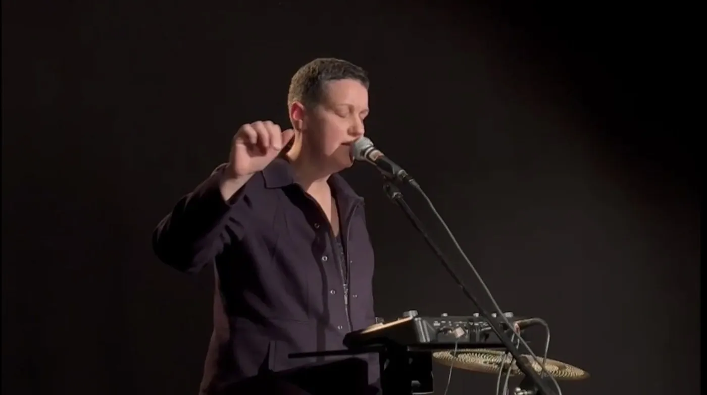 Marie-Laure Bourgeois chante au micro sur scène, une baguette à la main, devant ses percussions et ses machines.