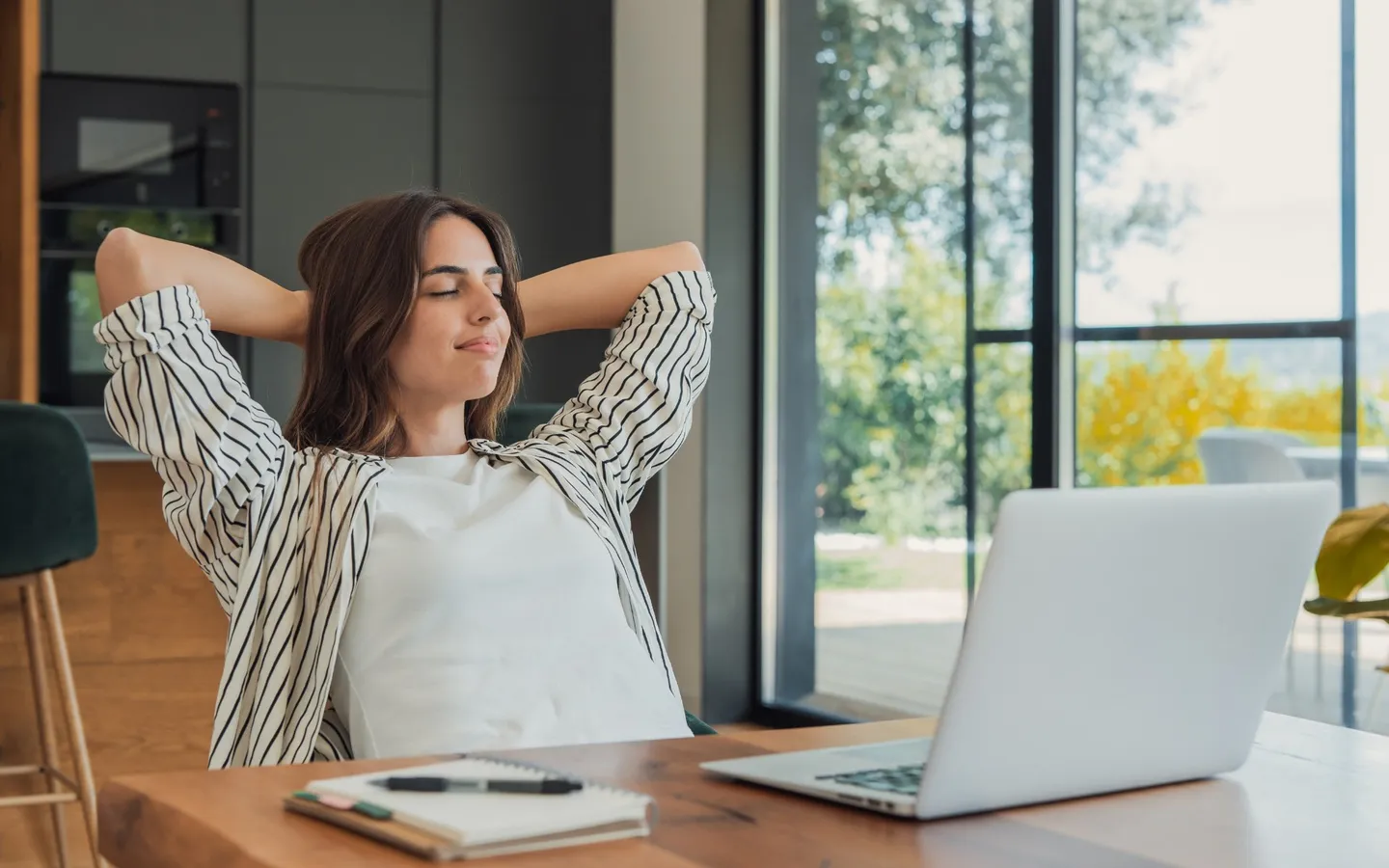 Une femme assise à son bureau, les yeux fermés et les bras derrière la tête, prend un temps de pause devant son ordinateur portable.