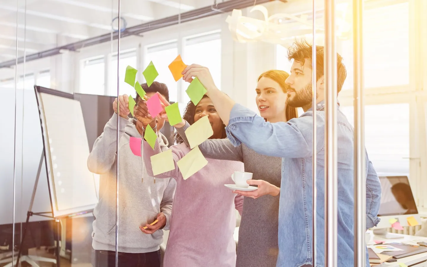 Trois collègues collent des Post-it de couleur sur une cloison vitrée, dans un bureau baigné de soleil.