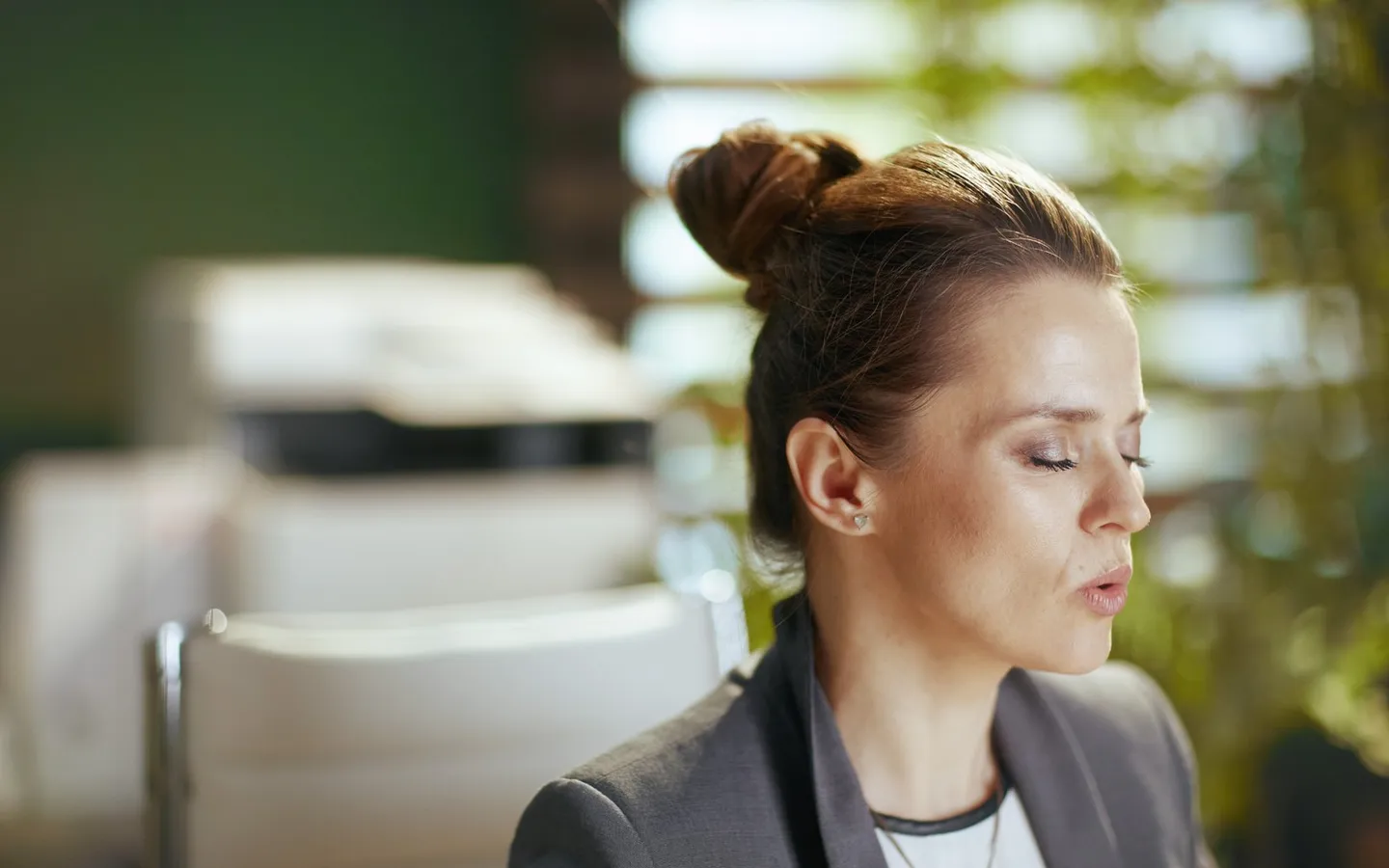 Une femme assise, les yeux fermés, en train d'expirer lentement, dans un bureau en arrière-plan flou.
