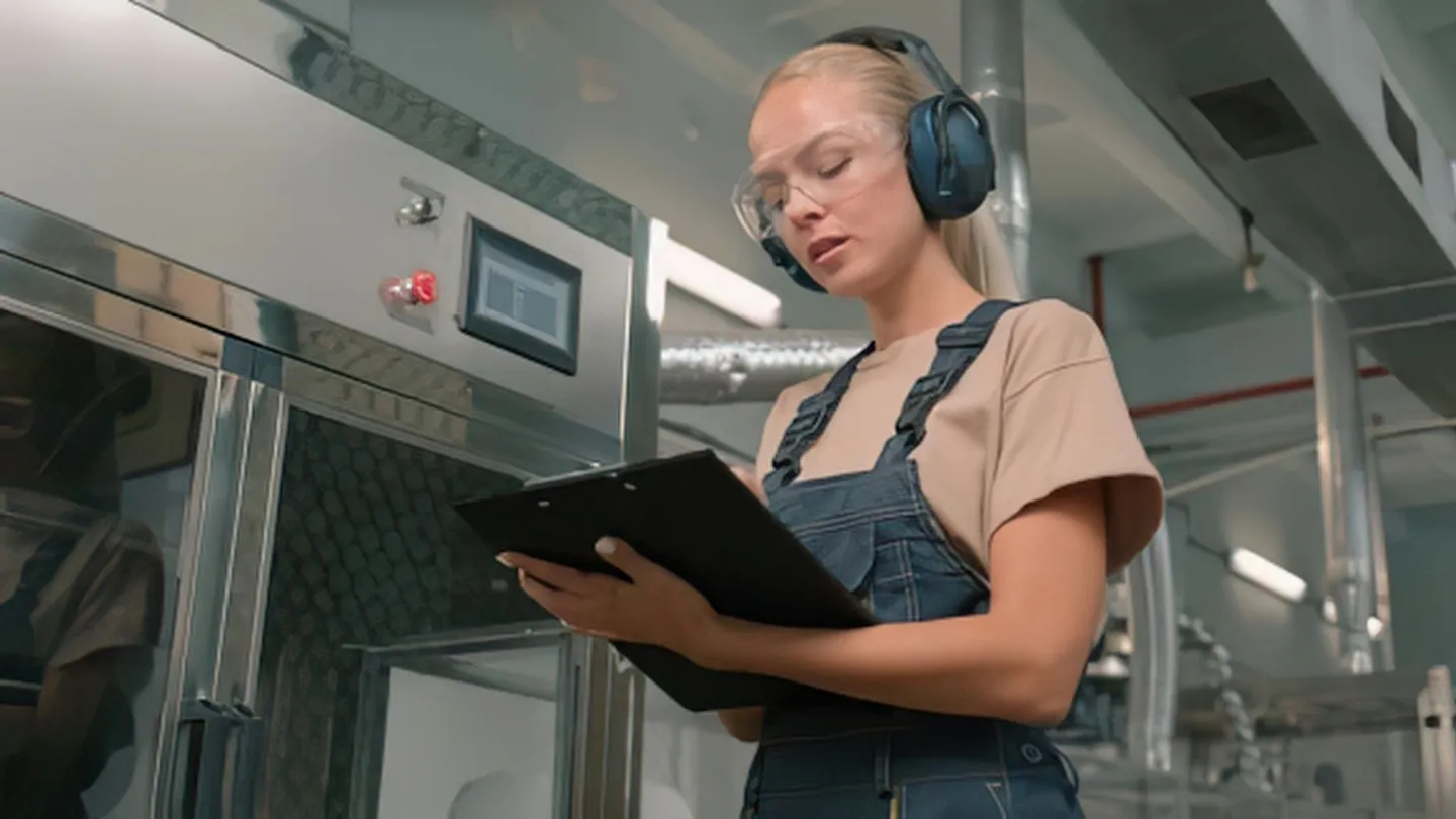Une technicienne en atelier industriel, casque audio sur les oreilles et lunettes de sécurité, consulte un porte-bloc.